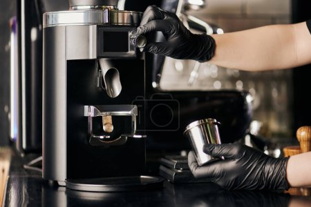 Photo for Barista in black latex gloves operating electric coffee grinder and holding metallic measuring cup - Royalty Free Image