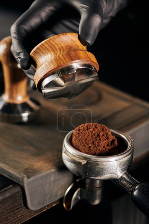 partial view of barista in black latex glove holding tamper near portafilter with ground coffee