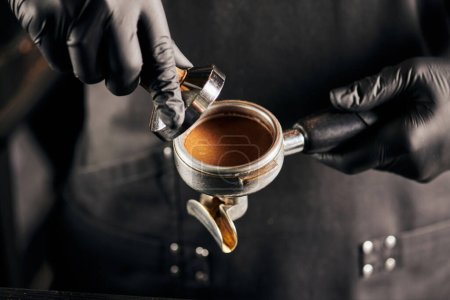 abgeschnittene Ansicht von Stampfer mit gemahlenem Kaffee und Portafilter in den Händen von Barista in schwarzen Handschuhen