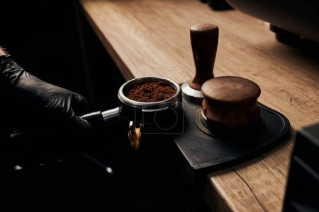 barista holding portafilter with grinded coffee, tamper, wooden table, cafe, alternative brew