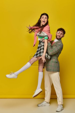 cheerful couple having fun, man in glasses lifting excited young woman on yellow backdrop, students