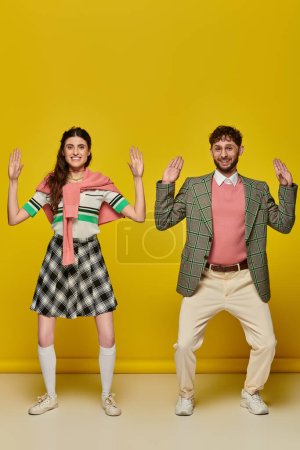 funny couple, happy young man and woman gesturing, standing on yellow backdrop, looking at camera