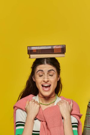 excited woman with closed eyes standing with book on head, yellow backdrop, student life, young