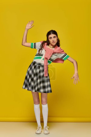 brunette woman acting like a doll, gesturing unnaturally, standing on yellow backdrop, student