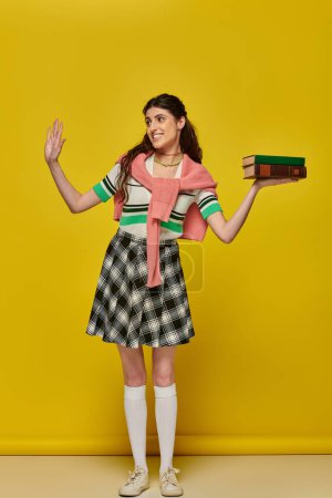 Photo for Happy student with books, brunette woman in checkered skirt waving hand on yellow backdrop, brunette - Royalty Free Image