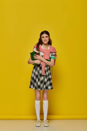 brunette student standing with books on yellow backdrop, young woman in skirt, college outfit