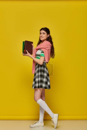 estudiante alegre de pie con libros sobre fondo amarillo, mujer joven en falda, traje de la universidad
