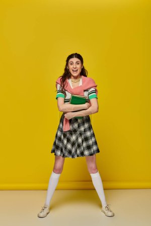 excited young woman in skirt standing with books on yellow backdrop, happy student, college outfit
