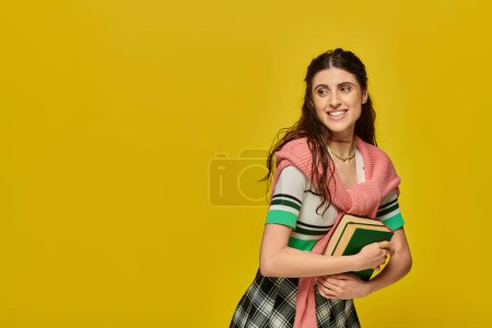 mujer joven positiva en falda de pie con libros sobre fondo amarillo, estudiante feliz, traje de la universidad