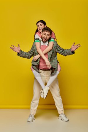Photo for Excited couple, funny, young man piggybacking brunette woman on yellow backdrop, student outfits - Royalty Free Image
