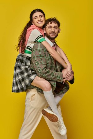 Photo for Couple, funny students, happy man piggybacking young woman on yellow backdrop, college outfits - Royalty Free Image