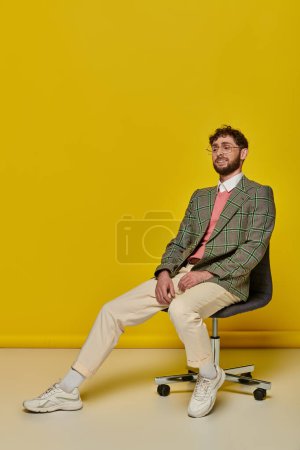 glücklicher bärtiger Mann sitzt auf Bürostuhl, gelber Hintergrund, Student im College-Outfit, Brille