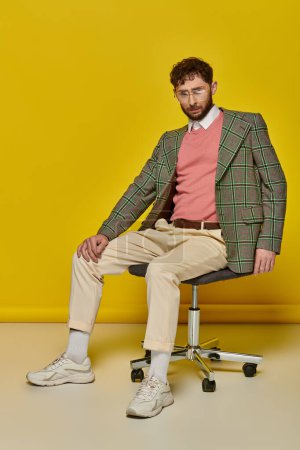 bärtiger Mann sitzt auf Bürostuhl, gelber Hintergrund, Student im College-Outfit und Brille