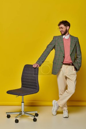 hombre positivo de pie con la mano en el bolsillo cerca de la silla de oficina, fondo amarillo, estudiante en gafas