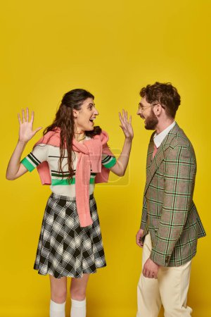 excited woman gesturing and looking at man on yellow backdrop, happy students, academic wear