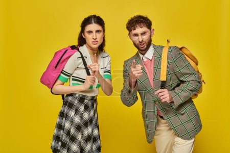Photo for Angry students standing with backpacks, looking at camera, pointing with finger, yellow backdrop - Royalty Free Image
