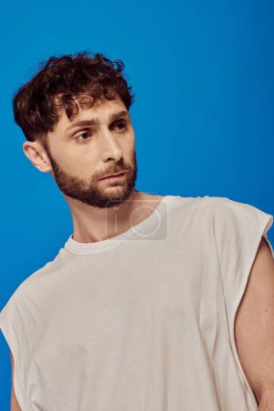 Photo for Bearded and curly man posing on blue background, white tank top, male fashion, looking away - Royalty Free Image