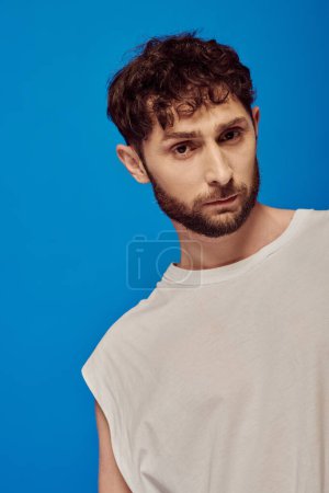 Photo for Bearded and curly man posing on blue background, white tank top, male fashion, looking at camera - Royalty Free Image