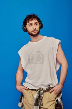 bearded man in wireless headphones listening music on blue background, white tank top, male fashion