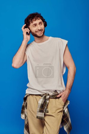 hombre alegre en auriculares inalámbricos escuchando música sobre fondo azul, mano en bolsillo, moda masculina