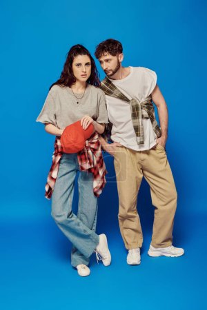 college couple posing in street wear on blue backdrop, woman with bold makeup, baseball cap