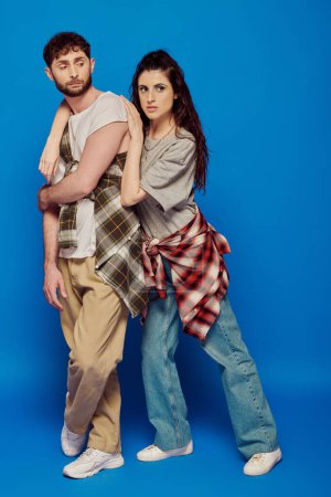 couple posing in street wear, blue backdrop, woman with bold makeup leaning on bearded man, trend