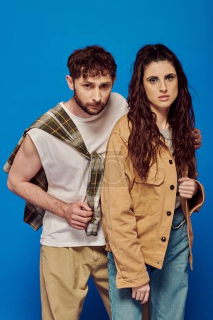 stylish couple in casual wear, blue backdrop, woman standing with bearded man, bold makeup, trends