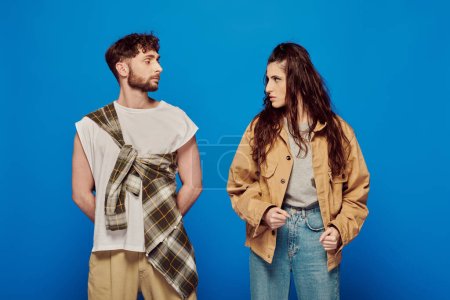 stylish couple in casual wear looking at each other, blue backdrop, woman standing with bearded man