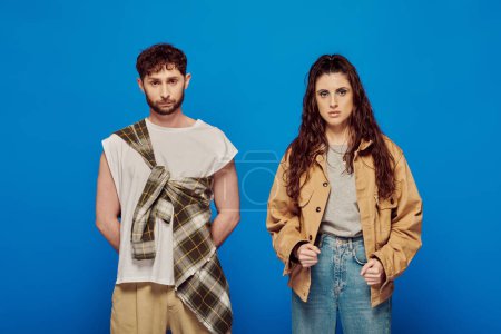 stylish couple in casual wear on blue backdrop, woman and man looking at camera, fashionable