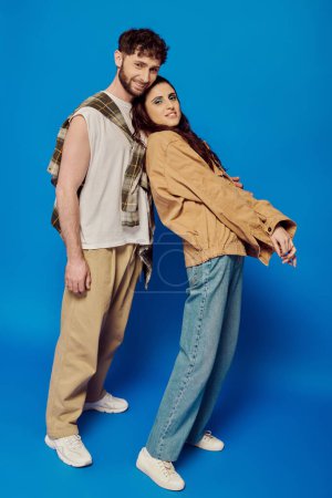 happy and stylish couple in casual wear posing on blue backdrop, woman and man looking at camera