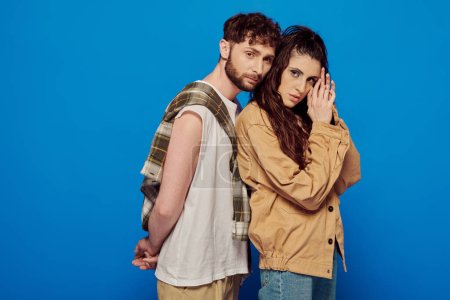 stylish couple in casual wear posing on blue backdrop, woman and man with beard looking at camera