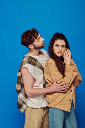 stylish couple in casual wear posing on blue backdrop, bearded man hugging woman, looking at camera
