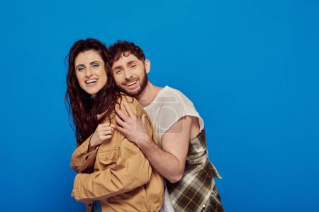 cheerful couple in casual wear posing on blue backdrop, bearded man hugging woman, bold makeup