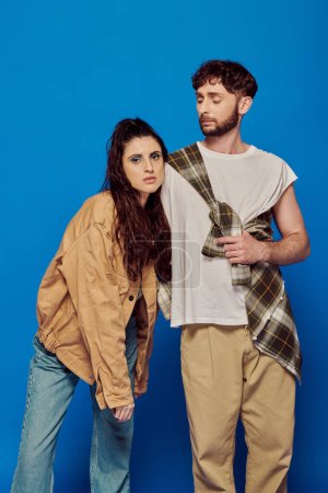 Photo for Fashionable couple in casual wear posing on blue backdrop, bearded man hugging woman, bold makeup - Royalty Free Image