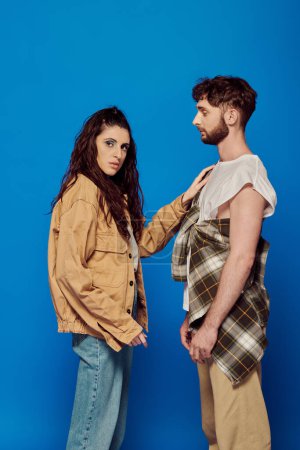 fashionable couple, casual attire, blue backdrop, woman touching chest of bearded man, bold makeup