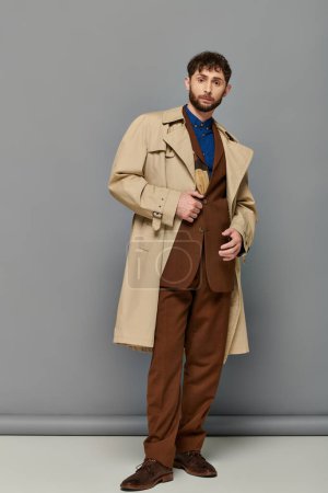 ropa de abrigo, hombre barbudo en gabardina posando sobre fondo gris, capas acogedoras, moda de otoño