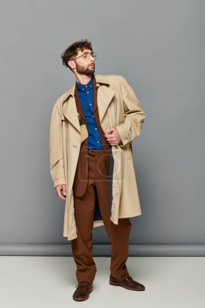 ropa de abrigo, hombre elegante en gafas y gabardina posando sobre fondo gris, capas acogedoras, moda de otoño
