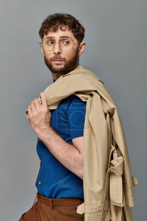 Foto de Hombre con estilo en gafas sosteniendo gabardina, fondo gris, modelo masculino mirando hacia otro lado, casual inteligente - Imagen libre de derechos