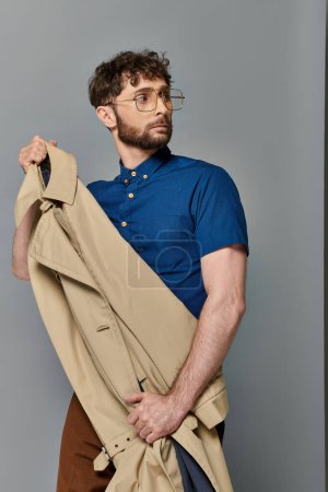 man in glasses holding trench coat, grey backdrop, stylish male model looking away, smart casual