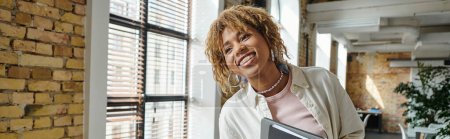 positive african american woman with braces smiling, holding folders in open space, startup, banner