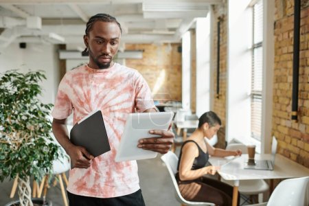 hombre afroamericano usando tableta, carpeta de retención, trabajando cerca de compañera de trabajo, proyecto de startup