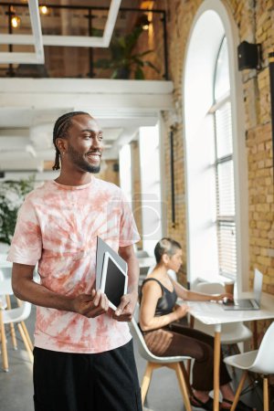 cheerful african american man holding tablet, folder, looking away near female coworker, startup
