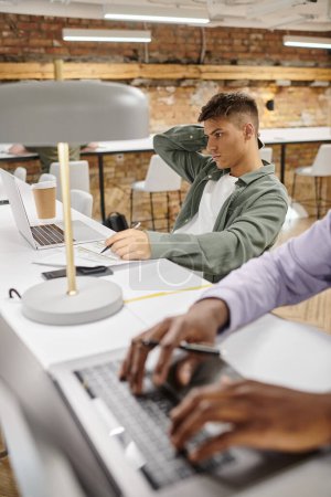 hombre concentrado usando el ordenador portátil, planificación de inicio, ideas, sentado cerca de un colega afroamericano