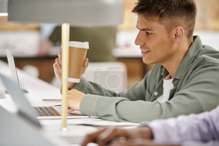 homme heureux tenant café pour aller et en utilisant ordinateur portable, environnement de bureau, coworking, projet de démarrage