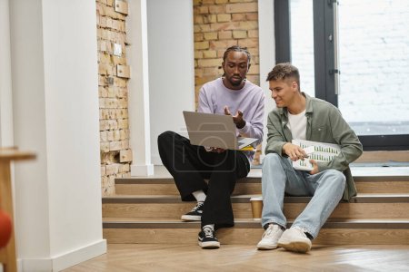 african american man showing project on laptop to male colleague, sitting on stairs, startup, modern