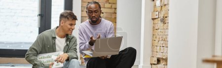 Afrikanisch-amerikanischer Mann zeigt männlichen Kollegen Projekt auf Laptop, sitzt auf Treppe, Start-up, Banner