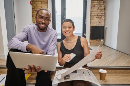happy man and woman holding gadgets and graphs, interracial colleagues looking at camera, startup