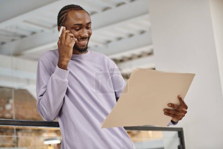 hombre afroamericano feliz mirando el proyecto en papel, hablando en smartphone, planificación, puesta en marcha
