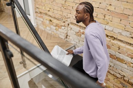 hombre afroamericano joven sosteniendo gráficos, caminando por las escaleras en coworking, gen z, startup, negocio