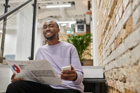positive african american man holding graphs and smartphone, sitting on stairs, coworking, startup